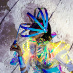 Catalyst Arts Bubble Performer Blowing Bubbles on a Large Manual Bubble maker while wearing a colorful costume and a creative headress